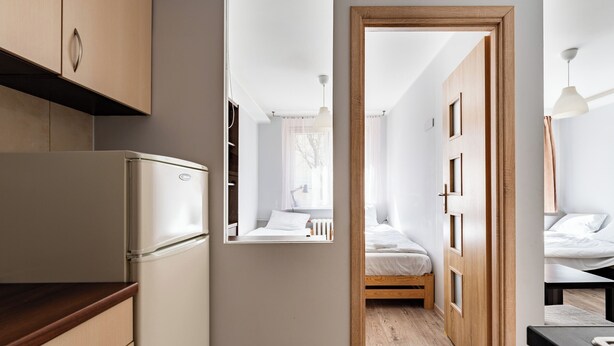 Un pasillo estrecho con una kitchenette a un lado, una cama con cabecera de madera y una mesita con lámpara.