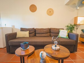 Een woonkamer met een bruine bank, twee ronde houten tafels, een kleine decoratieve wereldbol en een plafondventilator.