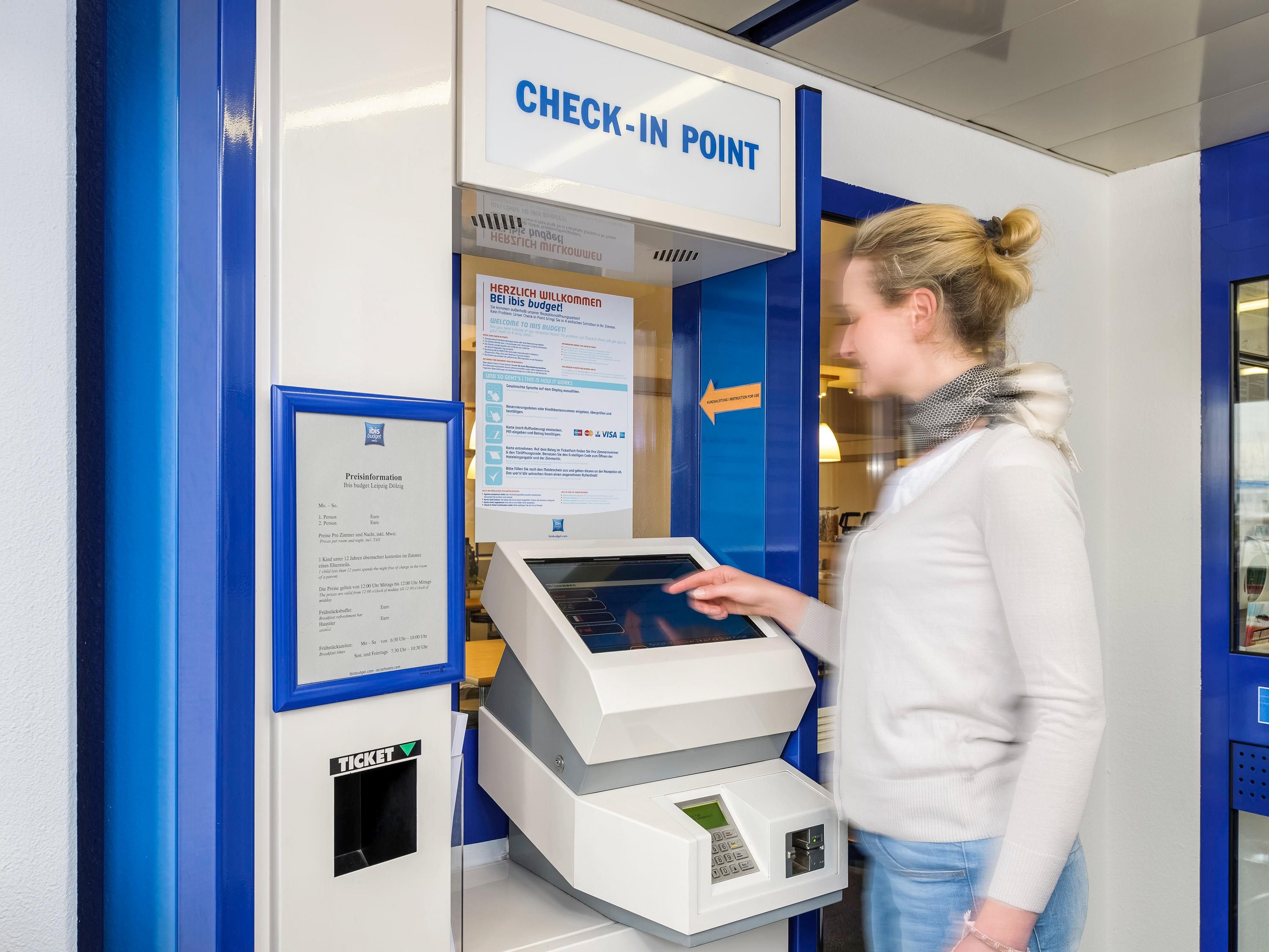 Un punto de registro con un quiosco digital, un cartel que dice 'CHECK-IN POINT' en inglés y una ranura para 'TICKET'.