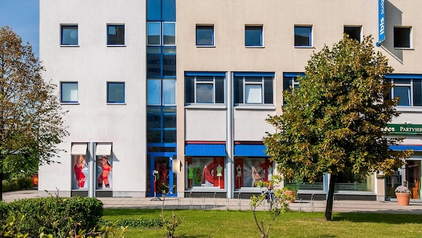 A multi-story hotel with a central glass elevator shaft, a blue awning over the entrance, and a visible sign for party services. - Ibis Budget Berlin Ost (Berlin)