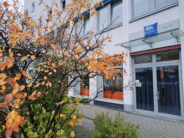 Entrada de un hotel con mostrador de recepción, un cartel de 'ibis budget hotels' y un edificio de fachada blanca.