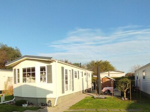 Une maison mobile de plain-pied, dotée d’un porche à l’avant, d’un petit jardin à l’arrière avec un arbre, et sous un ciel dégagé.