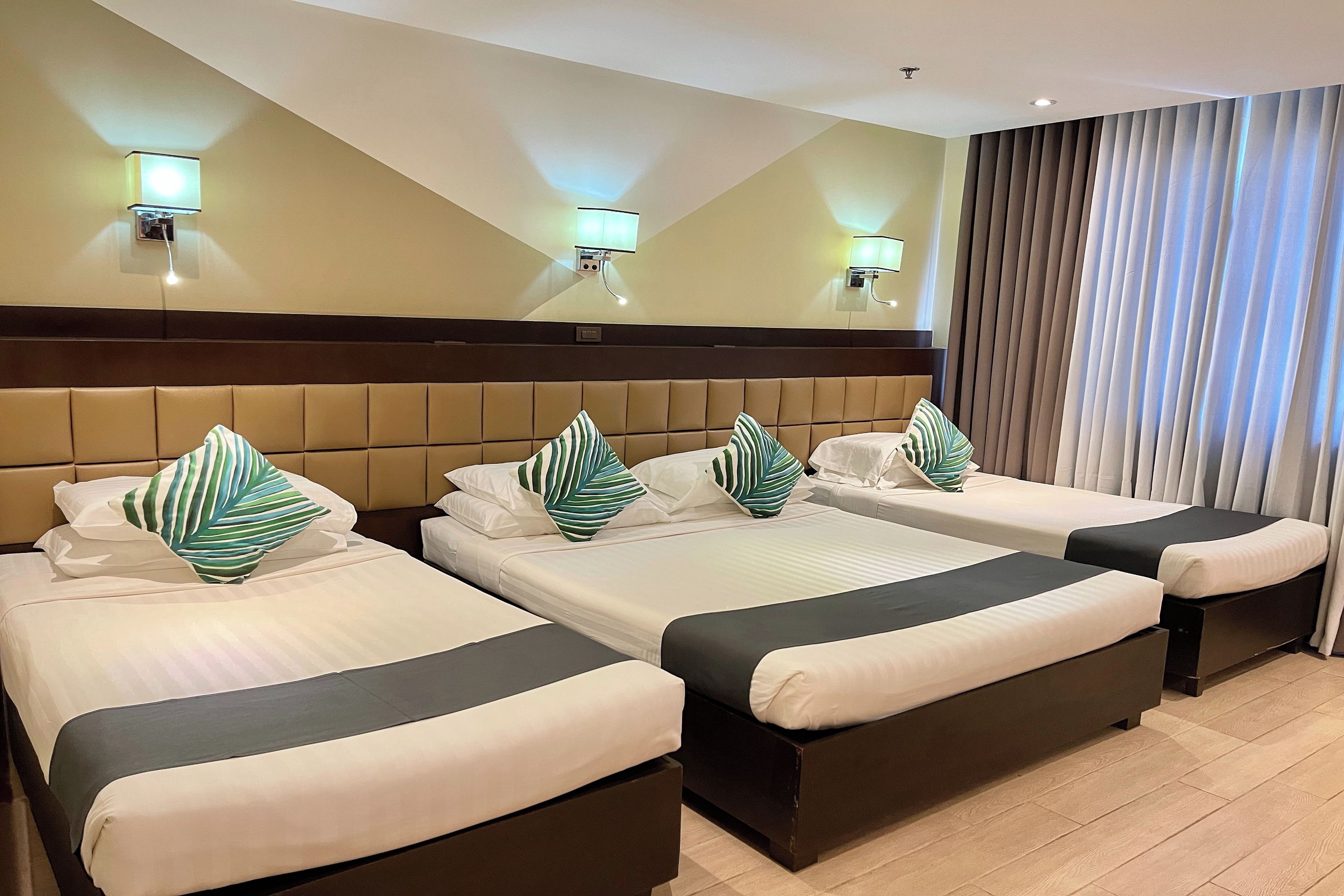 Habitación de hotel con tres camas, cabecera, luces fijadas a la pared y un ventanal con cortinas.