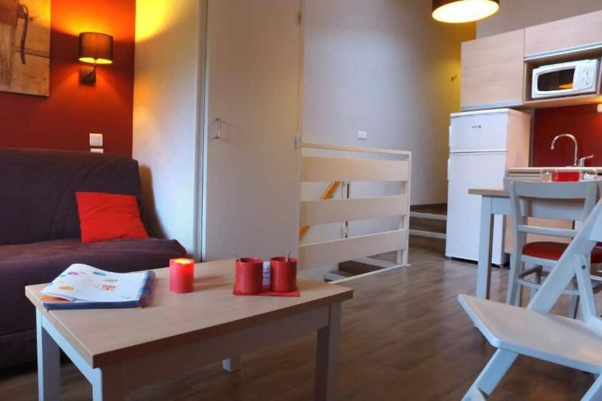 Un espace de vie compact comprenant un canapé, une petite table avec des livres et des bougies, un coin cuisine et un espace repas.