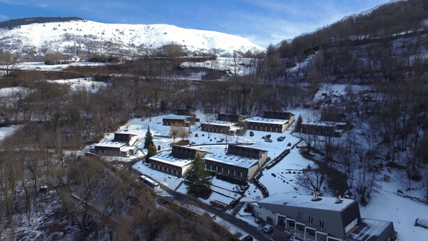 A snow-covered residential area with multiple two-story buildings, a central road, and a backdrop of snow-capped mountains.