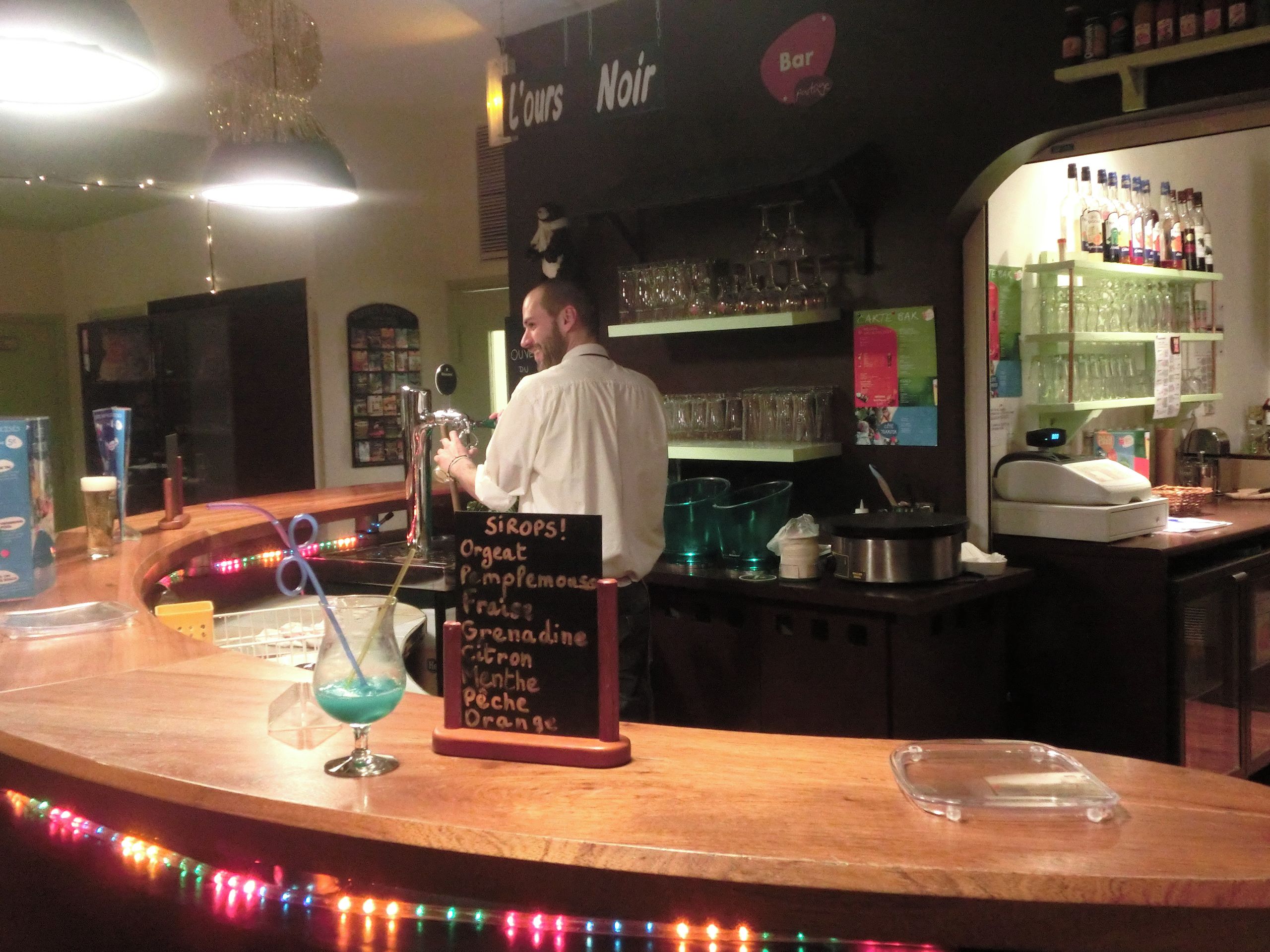 Eine Bar mit einem Barkeeper, einer Karte der angebotenen Getränke und Regalen mit Flaschen und Gläsern.