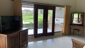 A room with a television on a wooden cabinet, a desk with a chair, and a sliding glass door leading to a balcony with a view of a green landscape.