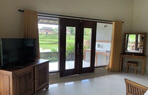A room with a television on a wooden cabinet, a desk with a chair, and a sliding glass door leading to a balcony with a view of a green landscape. - Bagiya Suites & Villas by Zuzu (Sebatu)