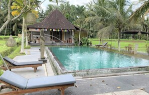 A pool with lounge chairs, a small pavilion, and lush greenery.