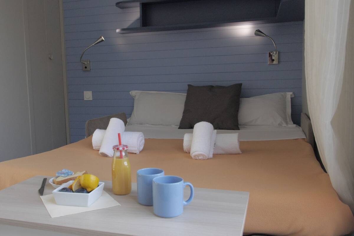 Una cama bien hecha con almohadas y toallas, una mesita de noche con un vaso de jugo, un cuenco de fruta y dos tazas.