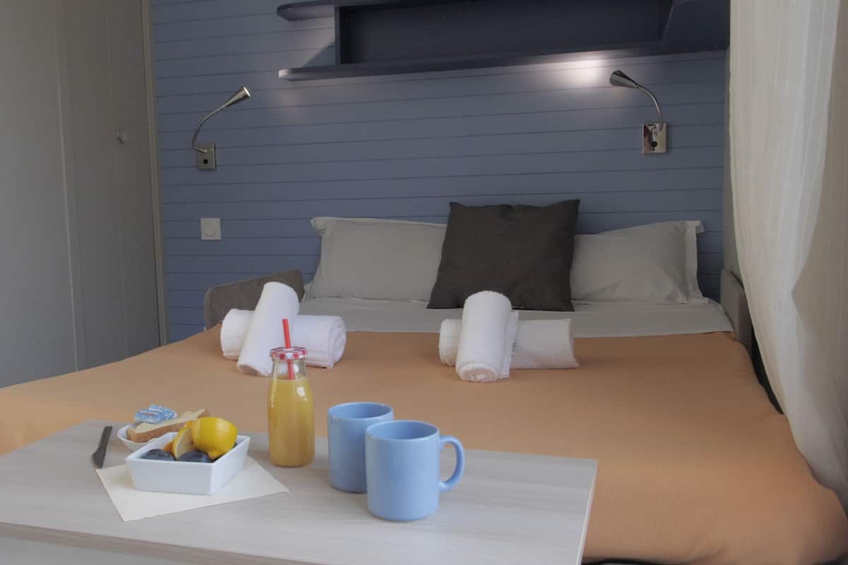 Un lit bien fait, avec des oreillers et des serviettes, une table de chevet sur laquelle se trouve un verre de jus de fruits, un bol de fruits et deux tasses.