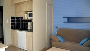 Eine kompakte Küche mit Geschirrspüler, Mikrowelle und schwarzem Fliesenrückwand. Im Wohnbereich steht ein braunes Sofa mit blauen Kissen.