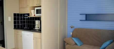 Cocina compacta con lavavajillas, microondas y salpicadero de azulejos negros. En la zona de estar, un sofá marrón con cojines azules.