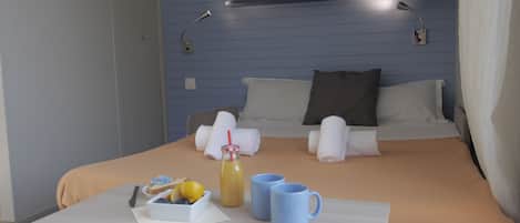 Una cama bien hecha con almohadas y toallas, una mesita de noche con un vaso de jugo, un cuenco de fruta y dos tazas.