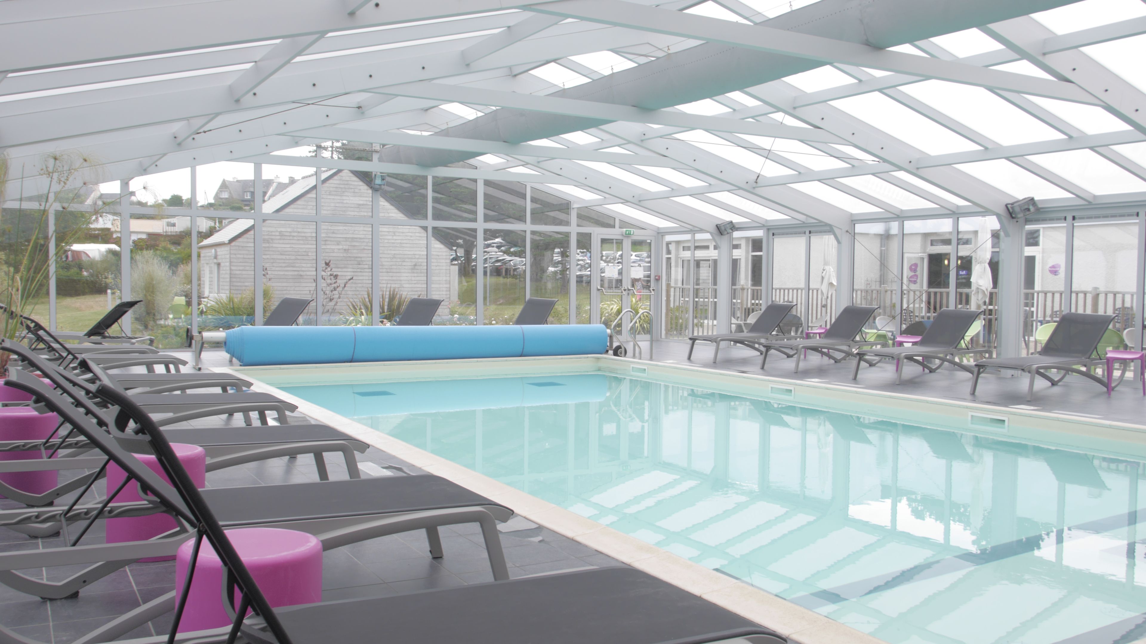 An indoor swimming pool with clear glass walls, lounge chairs, and a covered roof structure.