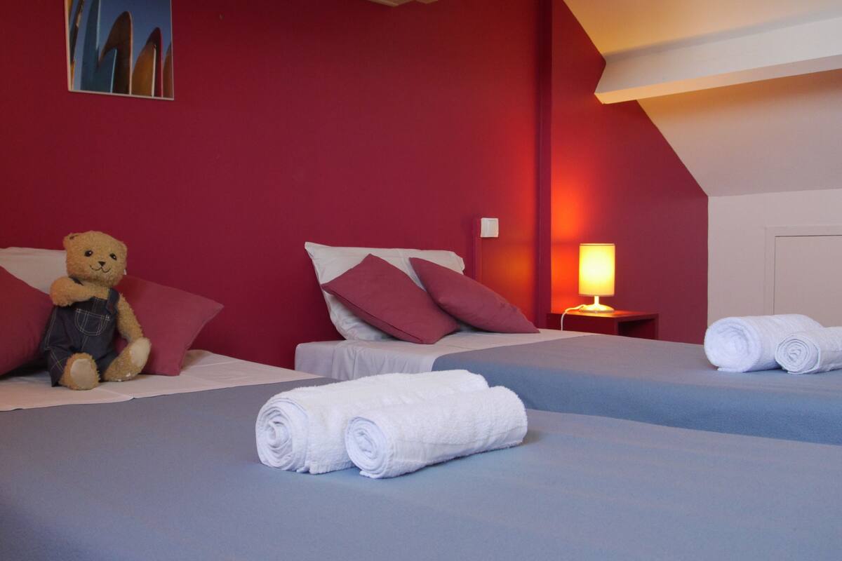 Habitación de hotel con dos camas, un oso de peluche en la cama de la izquierda y toallas blancas enrolladas en el suelo.