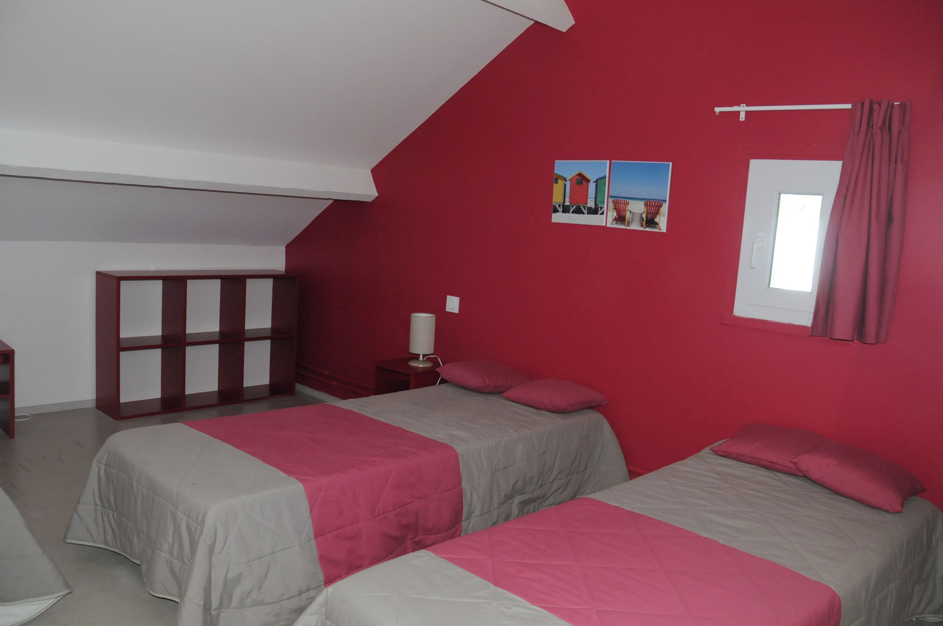A room with two beds, a red wall, a window with curtains, and a small table with a lamp.