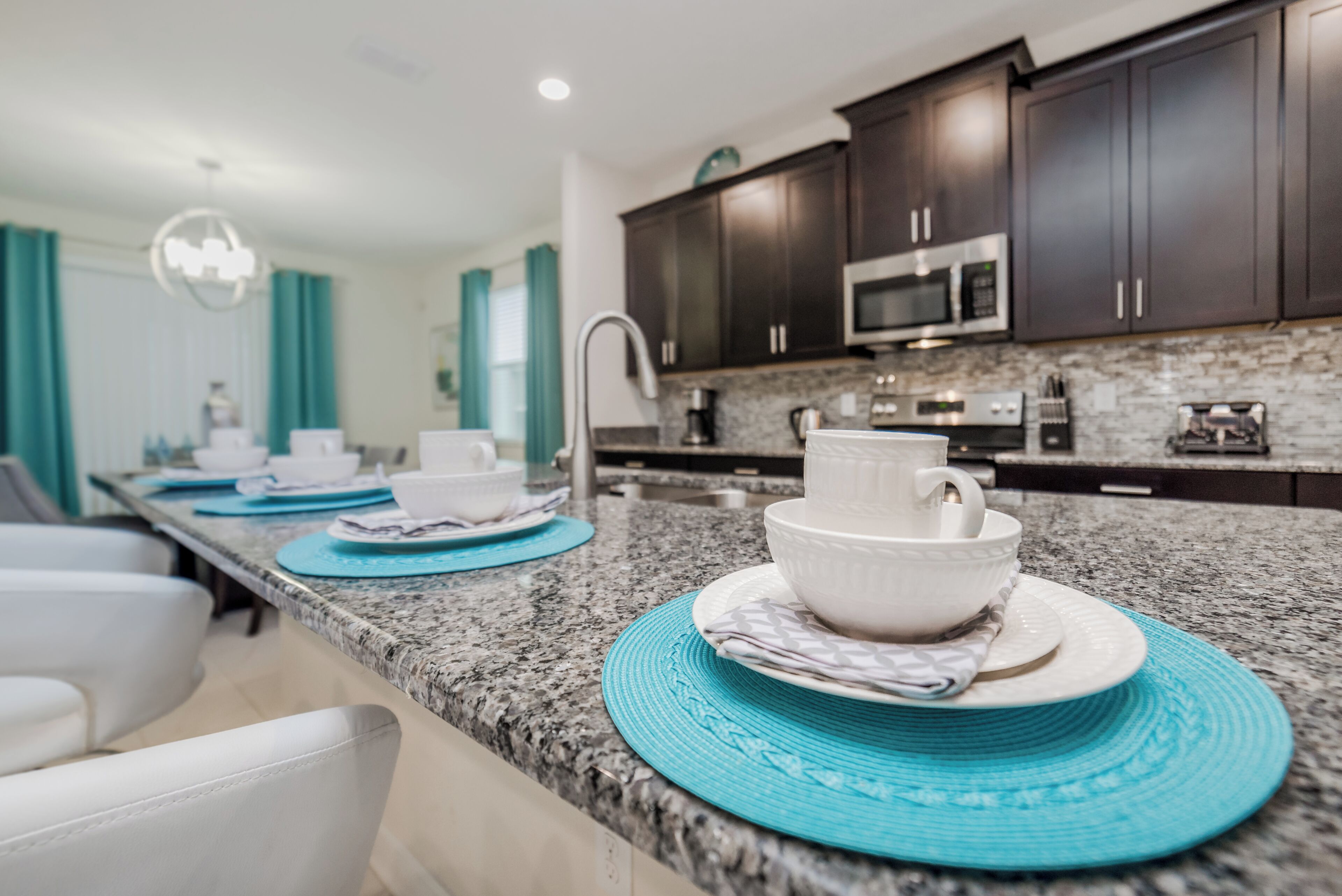 A modern kitchen with dark cabinets, a granite countertop, and a dining area with white chairs and blue placemats.