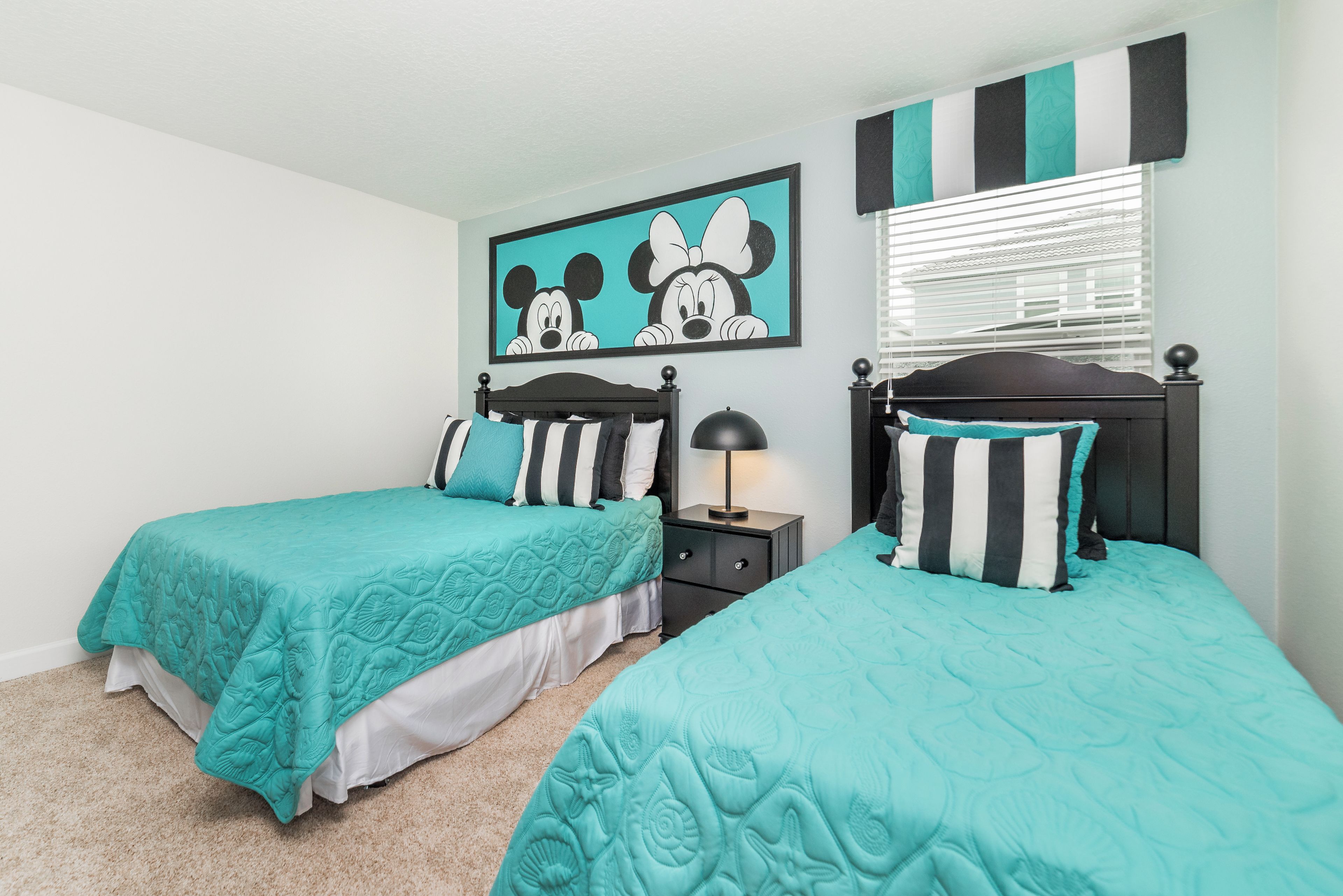 Une chambre à deux lits avec deux lits simples, une table de chevet, une lampe, un tableau de Mickey Mouse et des rideaux à rayures.