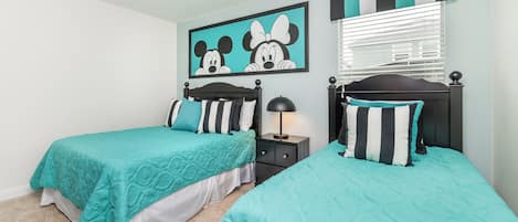 Habitación doble con dos camas, una mesita de noche, una lámpara, un cuadro de Mickey Mouse y cortinas a rayas.