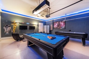 Uma mesa de bilhar com superfície de feltro azul, um projetor e um ventilador de teto em um ambiente decorado com tema de Star Wars.