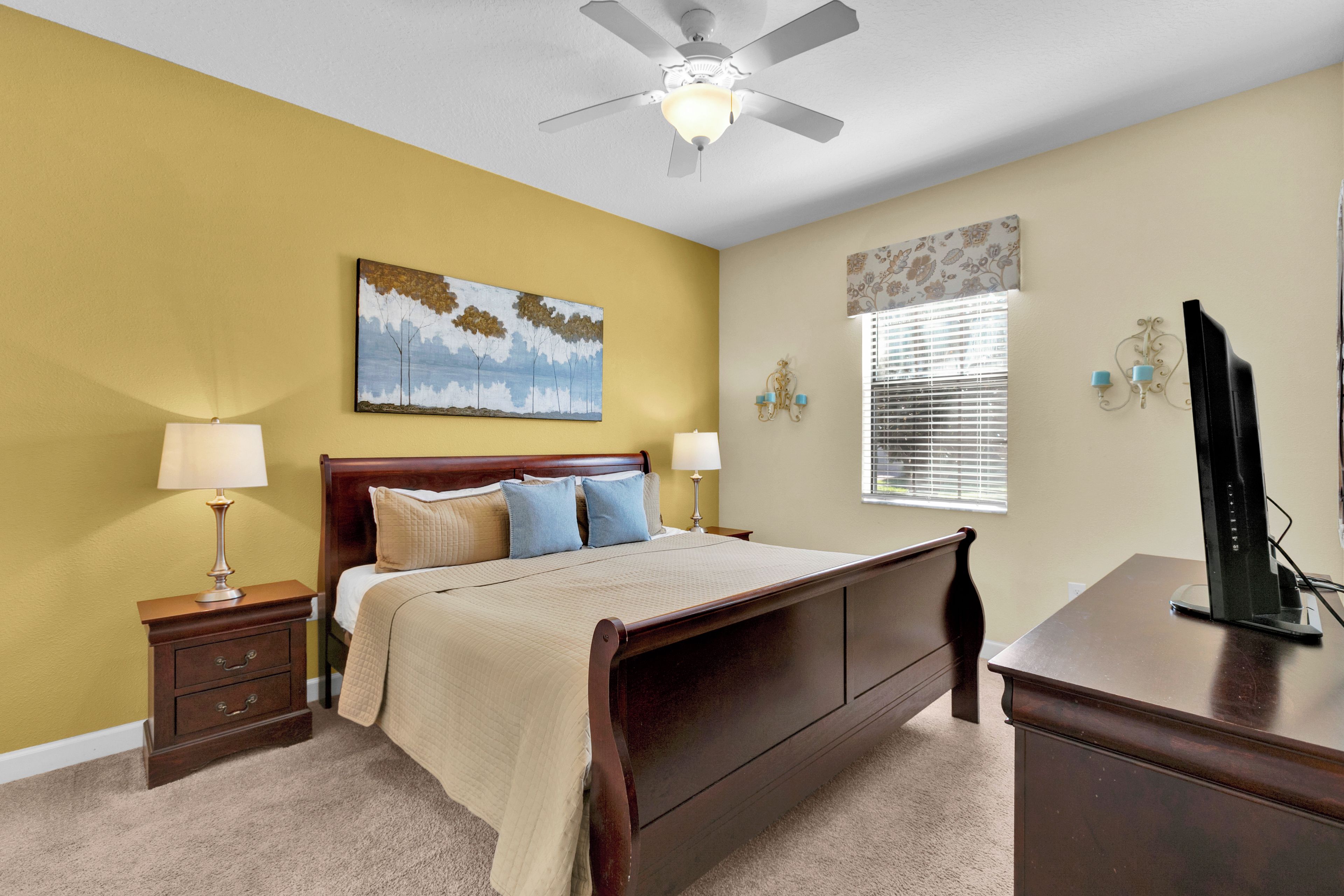A bedroom with a bed, nightstands, a desk, a ceiling fan, and artwork on the walls.