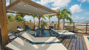 Un espace extérieur aménagé, abrité et doté de chaises longues et de tables, une pergola illuminée de guirlandes lumineuses, et une vue sur la plage et les bâtiments en arrière-plan.