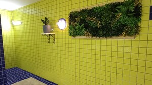 Une salle de bain avec des murs recouverts de carreaux jaunes, un sol carrelé de bleu, un tapis beige, une plante en pot et un luminaire fixé au mur.