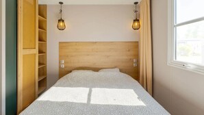 Une chambre à coucher avec une tête de lit en bois, deux lampes fixées au mur, une fenêtre avec des rideaux et une bibliothèque intégrée.