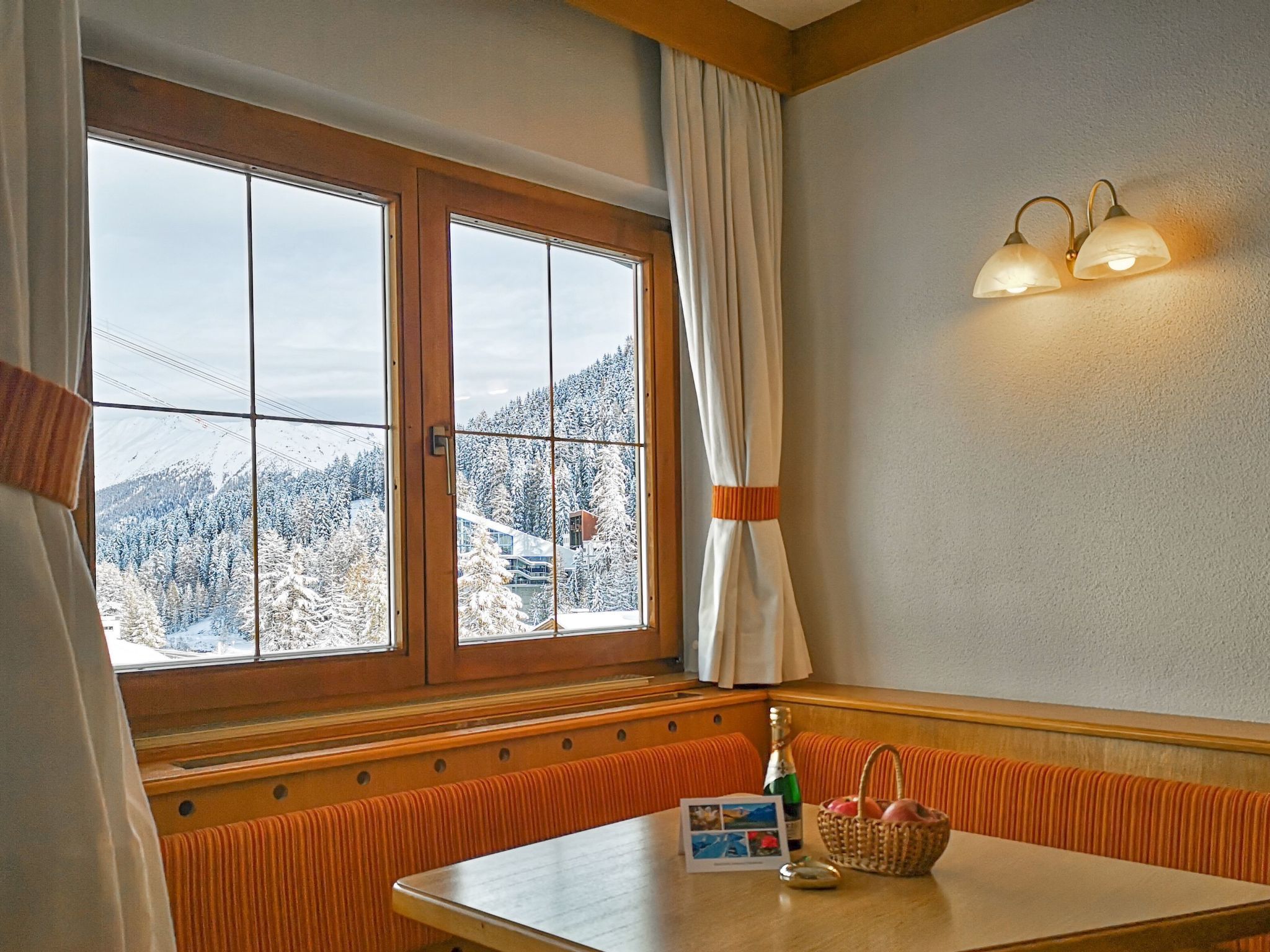 A hotel room with a large window offering a view of snow-covered mountains. There is a table with a basket and a bottle on it.