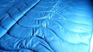 Een bed met een blauwe sprei en kussens, een houten hoofdbord en een klein nachtkastje met een lamp.
