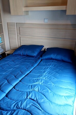 Una cama con colcha y almohadas azules, cabecera de madera y una mesita de noche con lámpara.