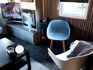 Ein modernes Wohnzimmer mit Fernseher, einem kleinen Tisch mit einer Kaffeetasse, einem Sessel und einem Fenster mit Jalousien.