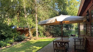 Eine Terrasse mit Tisch und Stühlen unter einem Sonnenschirm, umgeben von Bäumen und einem Holzzaun.