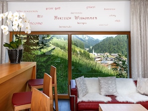 A room with a large window offering a view of a village and mountains, a wooden desk with a potted plant, a red sofa with patterned cushions, and a chair. - Apart Lischana, (Samnaun-Laret) by Interhome (Samnaun)