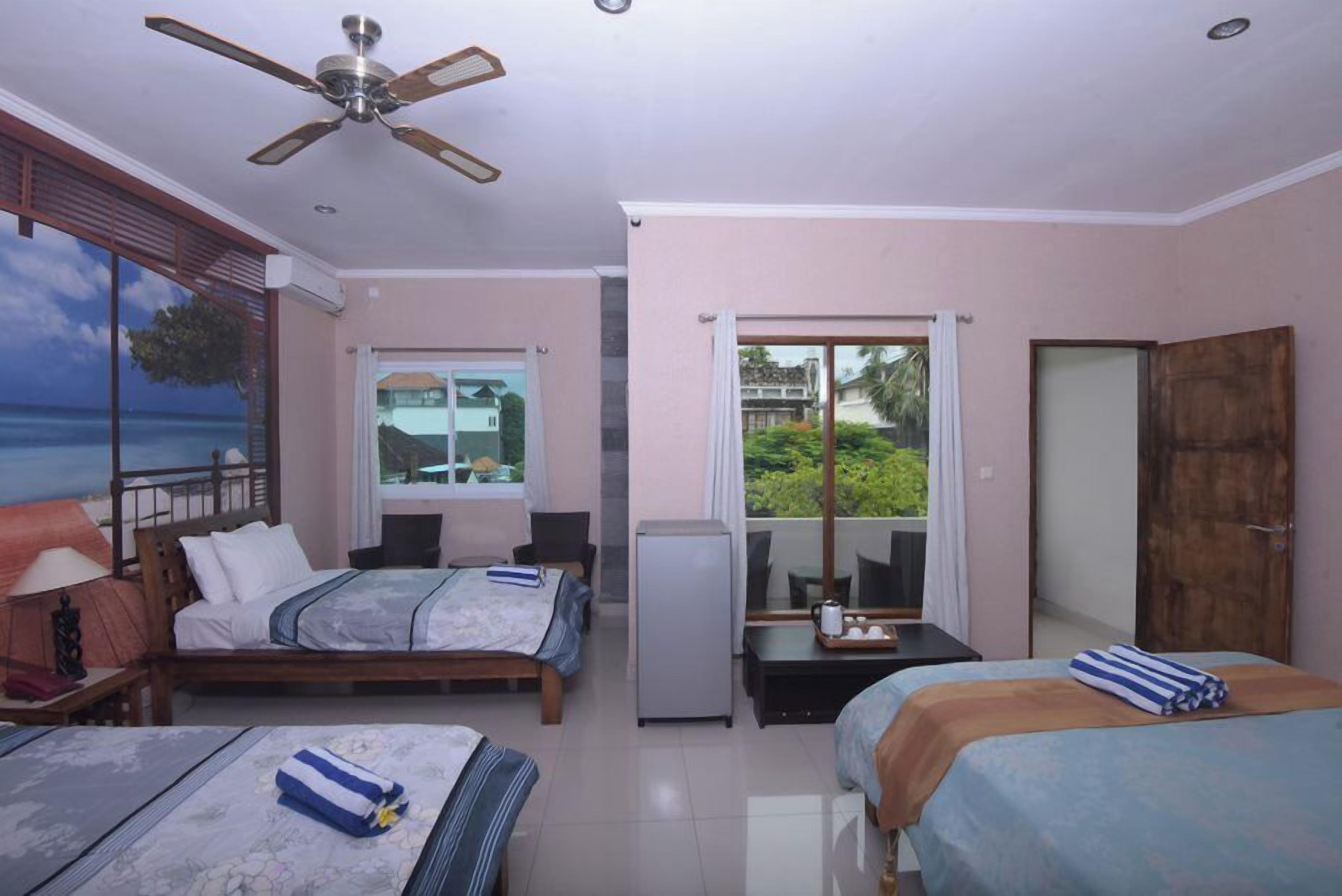 A hotel room with two beds, a ceiling fan, a refrigerator, a dining table, and a view of the ocean.
