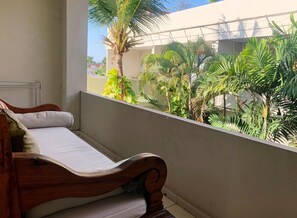 A balcony with a wooden bed, a view of a building, and lush greenery. - Su's Cottages Ii (Legian)