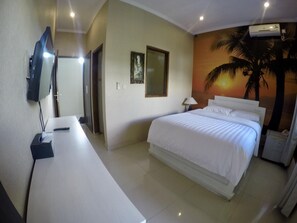 A hotel room with a bed, a desk, a TV, a painting, and an air conditioning unit. - Su's Cottages Ii (Legian)