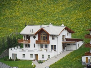 Ein zweistöckiges Gebäude mit Balkon und einer Treppe, die zum Eingang führt, vor einer Hügelkulisse.