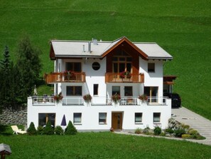 A two-story building with a balcony on the second floor, a stone wall, and a paved driveway. - Apartment MEZPRA142, (Samnaun-Compatsch) by Interhome (Samnaun)