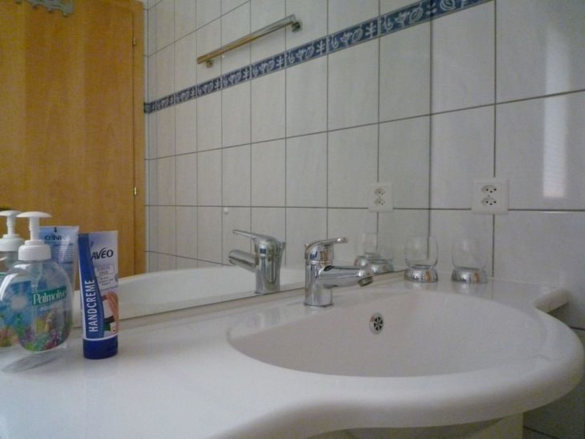 Een badkamer met een witte wastafel, twee kraanwerk en een spiegel. Op de wastafel staan twee flesjes handcrème.