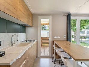 Eine moderne Küche mit Marmorarbeitsplatte, Edelstahlspüle und Holzschränken. Ein Holztisch mit Barhocker und Blick ins Freie durch eine Glastür.