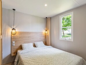 Ein Schlafzimmer mit Holz Kopfteil, zwei Nachttischlampen, einem Fenster mit Blick auf Grünflächen und einem Bett mit gemusterten Betttextilien.