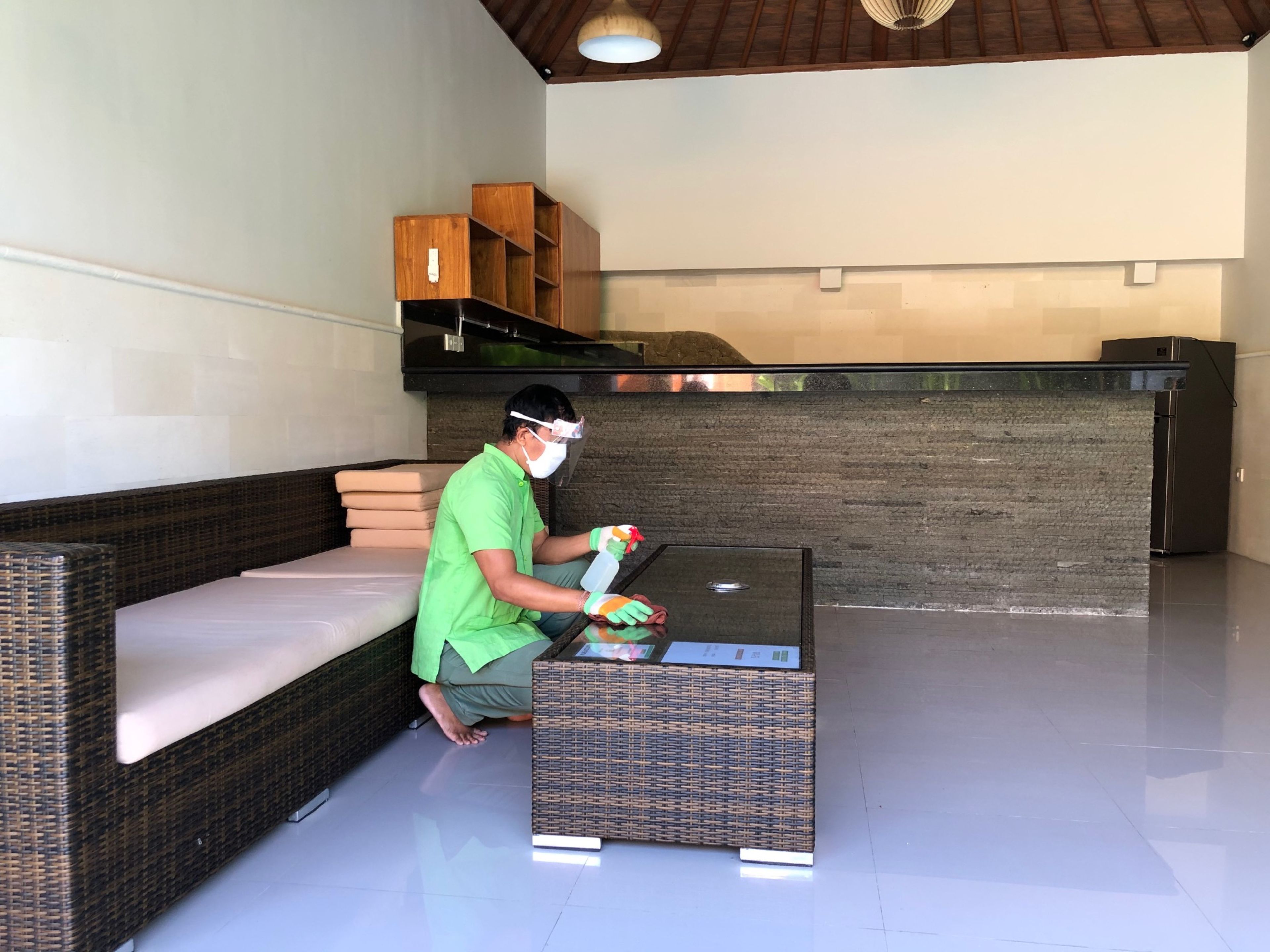 A man cleaning a glass table in a room with wicker furniture, a stone wall, and a ceiling light fixture.