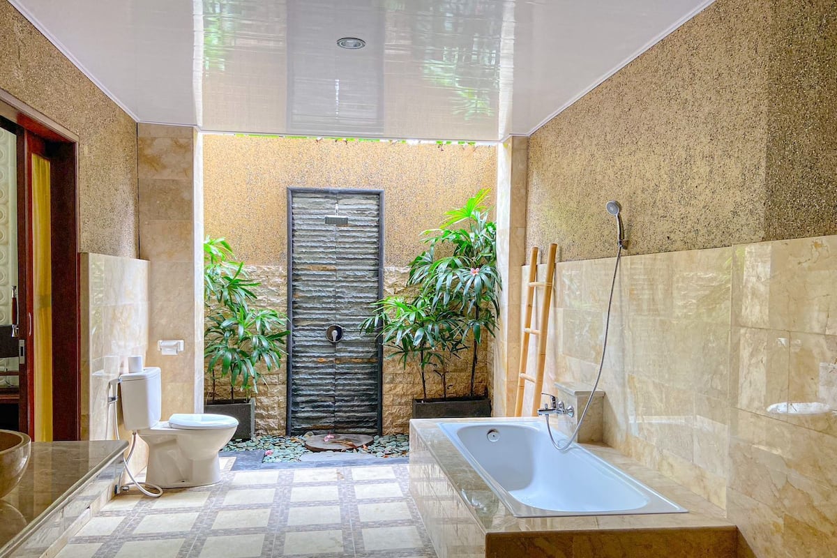 A modern bathroom with a bathtub, toilet, and sink. There are plants and a shower area.