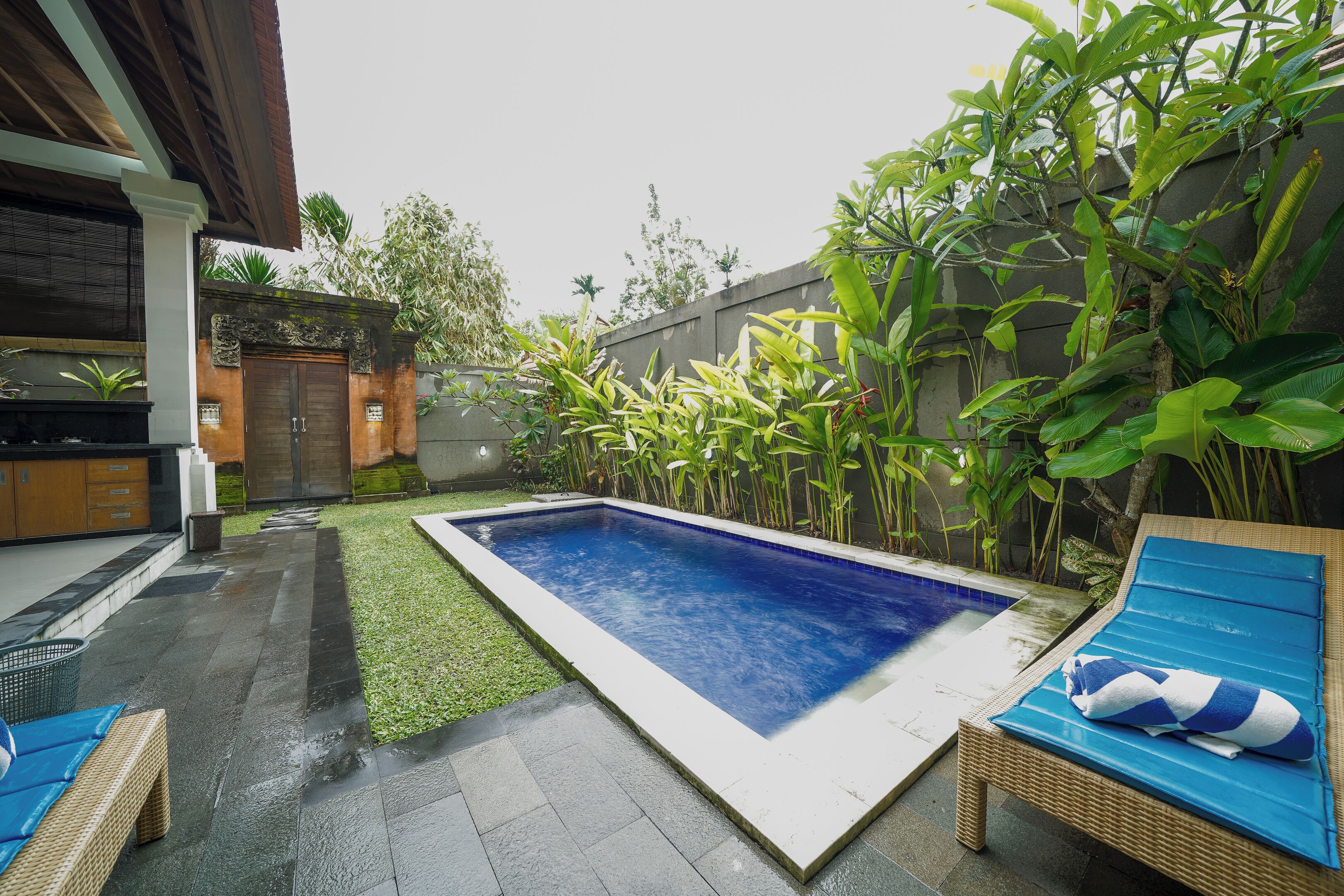 A rectangular swimming pool with a blue interior, surrounded by a small grassy area and various plants. There is a wicker lounge chair with a blue cushion and a striped towel on it.