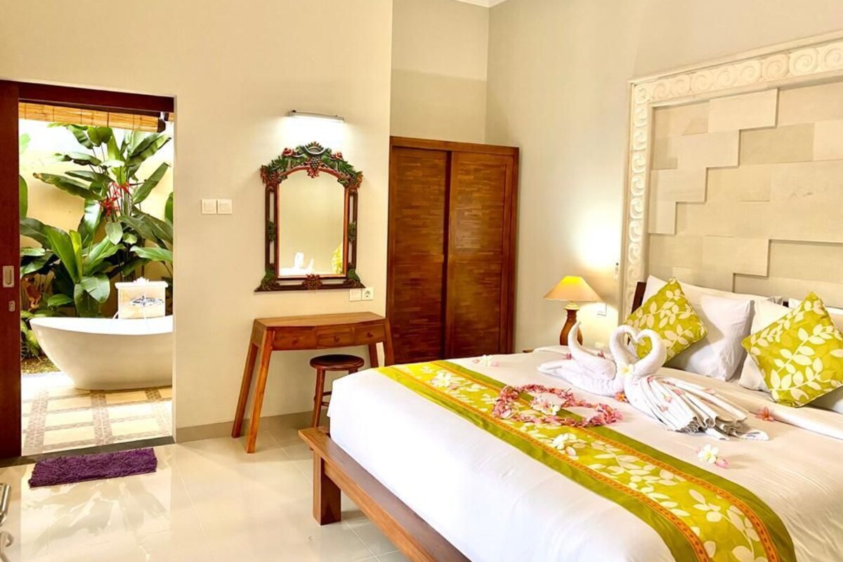 A hotel room with a bed, a desk, a mirror, a wardrobe, and a bathroom with a bathtub and potted plant.