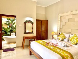 A hotel room with a bed, a desk, a mirror, a wardrobe, and a bathroom with a bathtub and potted plant. - Three Brothers Bungalows and Villas (Legian)