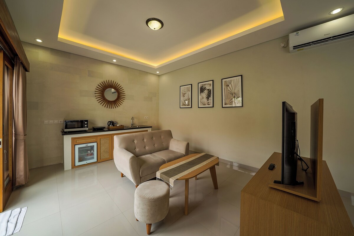 A modern living room with a sofa, ottoman, TV, and a kitchen area in the background.