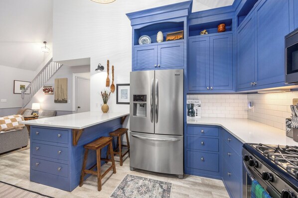 A modern kitchen with blue cabinets, a stainless steel refrigerator, a gas stove, and a white island with stools.